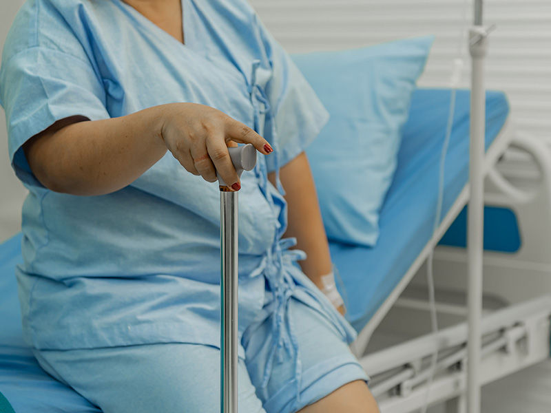 Adipositaskranke Patientin im mittleren Alter in blauer Krankenhauskleidung, die auf der Bettkante sitzt (blaue Bettdecke). Die rechte Hand hält einen Wanderstock. An ihrem linken Unterarm befindet sich ein weißes Pflaster. Eine Infusion steht neben dem Bett.
