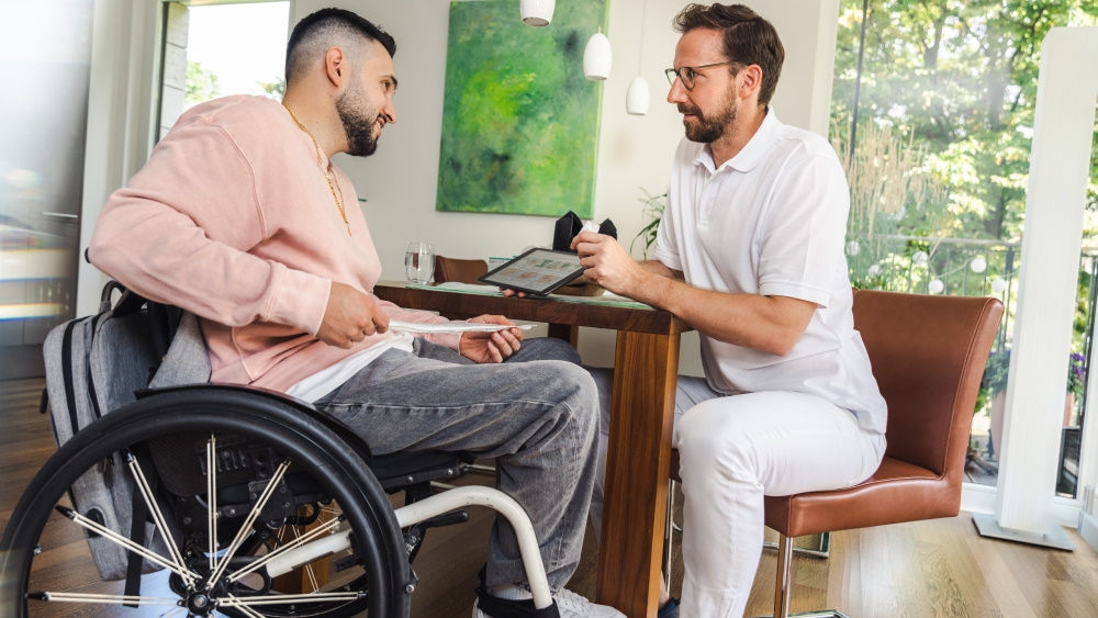 Ein Mann sitzt in einem Rollstuhl und hält einen Katheter in den Händen. Vor ihm gibt ein Krankenhausmediziner Anweisungen über ein Tablet