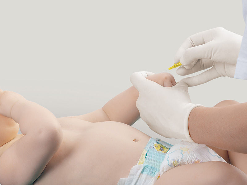 Baby liegt in Windeln. Der linke Arm wird mit einer behandschuhten Hand am Handgelenk gehalten. Eine behandschuhte rechte Hand hält eine gelbe Introcan Safety® Kanüle. Die Spitze der Kanüle zeigt zur Punktionsstelle.