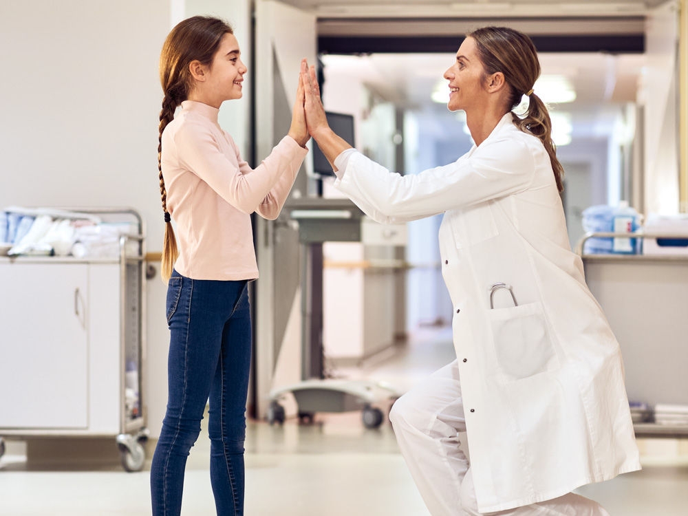 Ein dunkelhaariges Mädchen und ein dunkelhaariger Arzt stehen lächelnd einander in einem Krankenhauskorridor gegenüber. Der Arzt hat sich hingelegt, um sich auf Augenhöhe mit dem kleinen Patienten zu befinden. Sie schlagen einander mit beiden Händen hoch.
