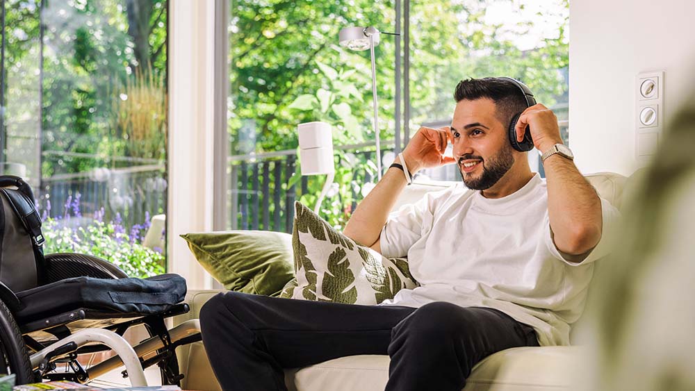 Rechts sitzt ein Mann mit Kopfhörern auf dem Sofa, er hält seine Hände an den Kopfhörern. Links ist ein Rollstuhl