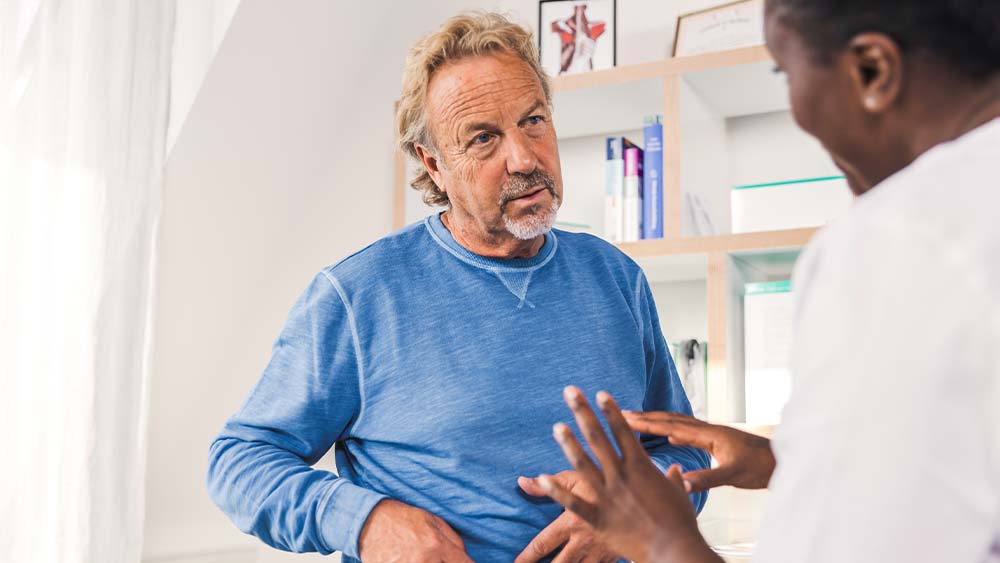Im rechten Hintergrund eine Ärztin, die mit den Händen gestikuliert und im Vordergrund ein Mann, der ihr zuhört