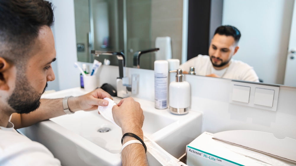 Ein Mann sitzt vor einem Spiegel in einem privaten Badezimmer und hält einen Katheter in den Händen.