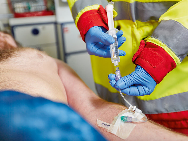Patient mit blauer Decke von der Bauchmitte aus bedeckt, liegt in einem Krankenwagen. Der linke Unterarm des Patienten wird mit einer grünen Introcan Safety® 3-Kanüle punktiert. Caresite-Ventil mit Infusionsleitung und weißer Schiebeklemme ist angeschlossen. Fixierung mit Kanülenpflaster. Person in Rettungsjacke und blauen Handschuhen hält gefüllte Spritze in der rechten Hand. Die linke Hand hält die Leitung unter dem Ventil.