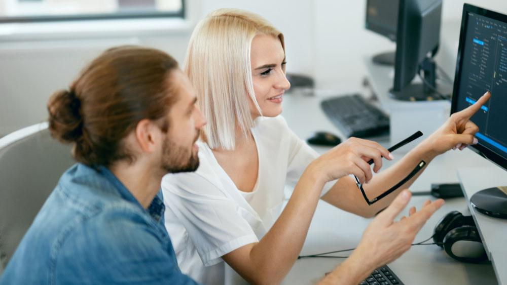 Zwei Pflegekräfte sitzen vor einem PC. Sie zeigt ihm etwas auf dem Bildschirm.