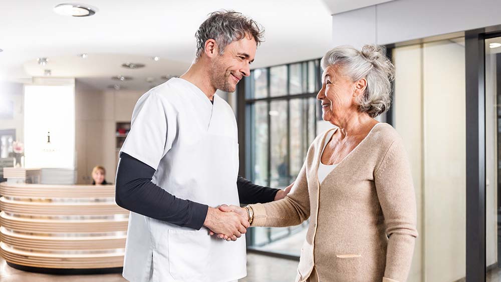 Ein Mann und eine ältere Frau schütteln sich die Hände in einer Krankenhausumgebung und symbolisieren Unterstützung und Verbundenheit während eines Besuchs.