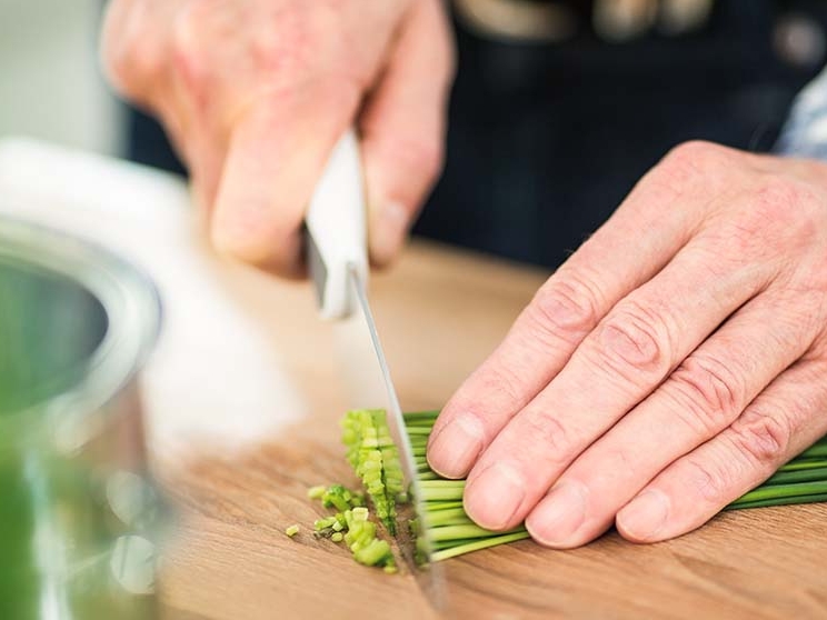 Eine Person schneidet gekonnt grüne Zwiebeln auf einem Schneidebrett aus Holz in einer gut beleuchteten Küche.