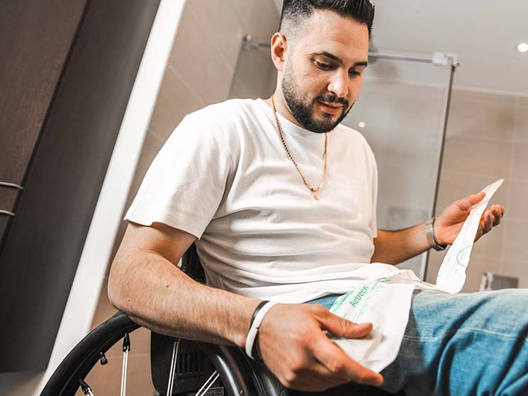 Ein Mann im Rollstuhl, ein Badezimmer im Hintergrund, hält einen Katheter in der Hand