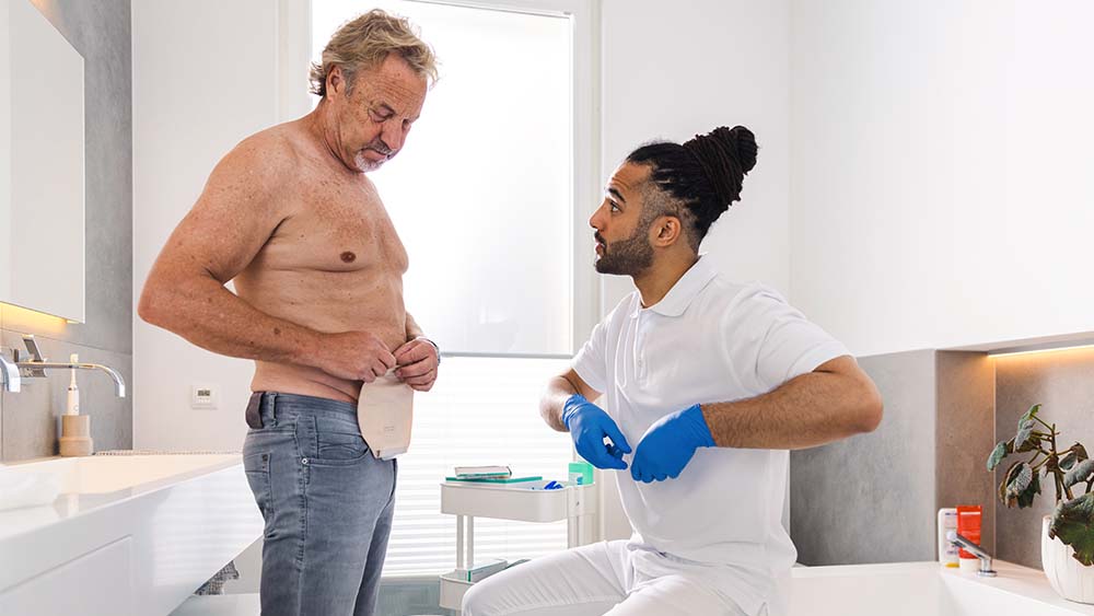Mann mit nacktem Oberkörper in einem privaten Badezimmer manipuliert einen Stomabeutel auf dem Bauch. Krankenschwester sitzt auf dem Badewannenrand und gibt Anweisungen.