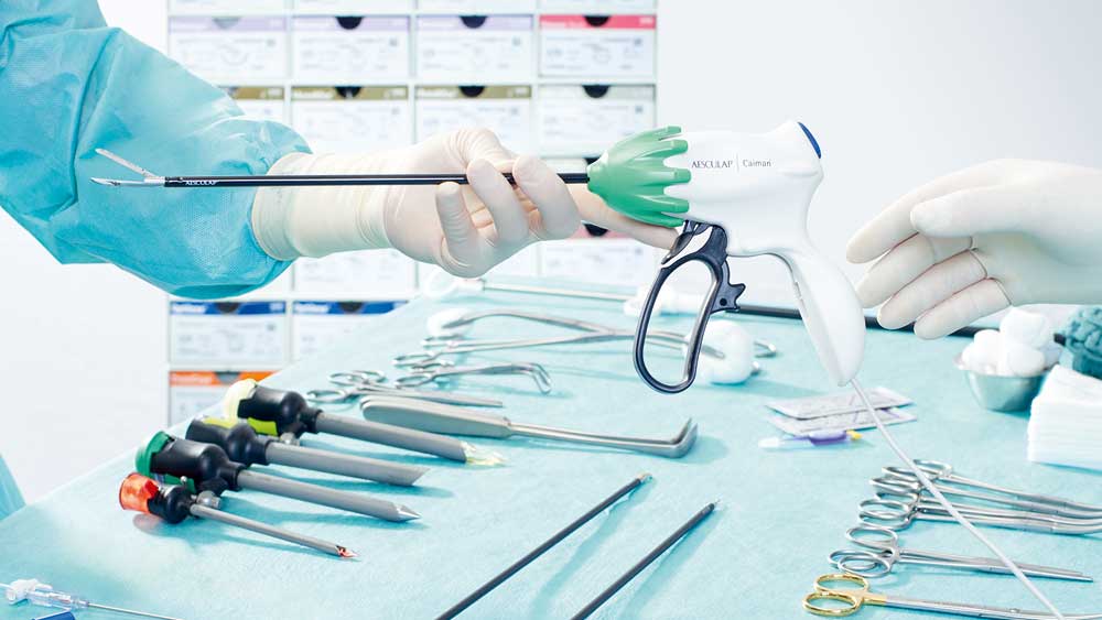 Ein Detail einer minimalinvasiven Operationssituation: Eine Person in OP-Kleidung übergibt einem anderen einen Caiman; im Hintergrund steht ein Tisch mit übersichtlich angeordneten chirurgischen Instrumenten für eine Operation